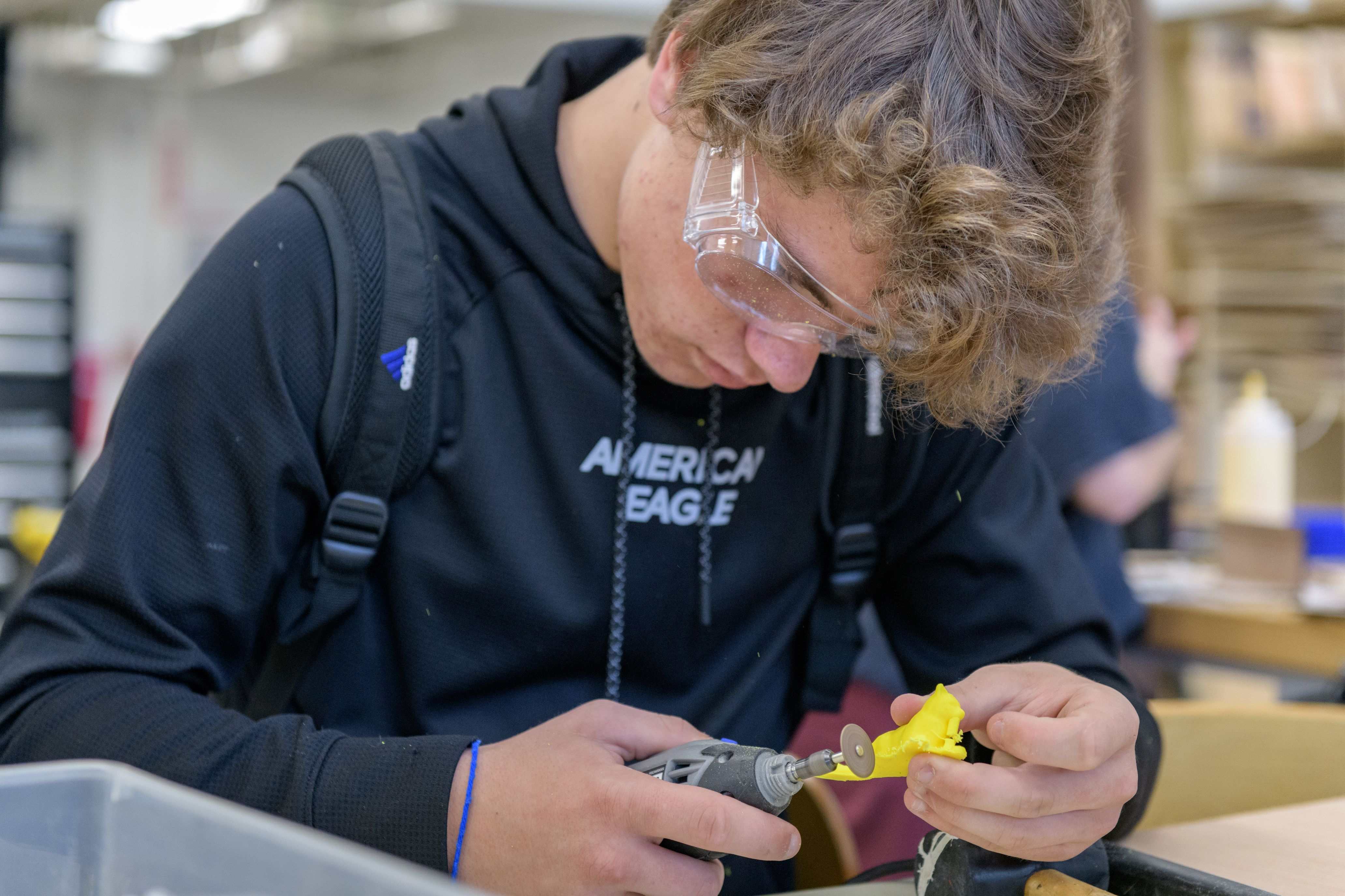 Pre-college student in safety glasses uses a rotary tool on a small yellow object as a part of his MakerSpace course