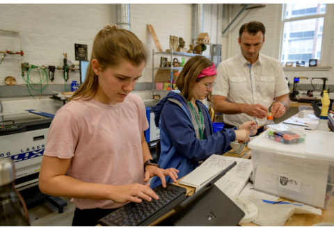 A Pre-College types on a keyboard in a workshop, with another student receives support from instructor behind her.