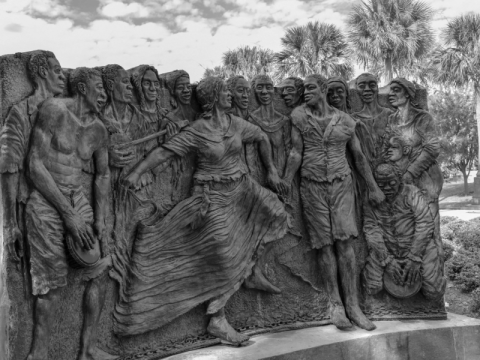 Dark stone bas-relief sculpture of numerous figures; a woman in a flowing dress leads a group.