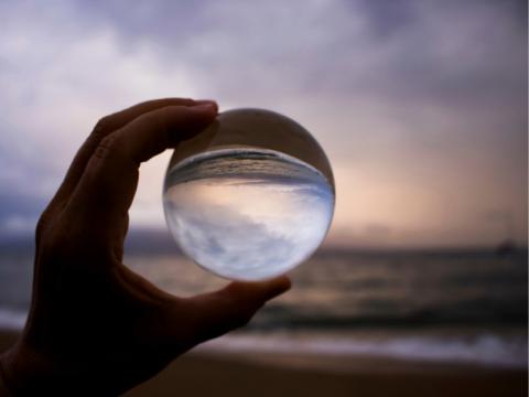 Hand holds a crystal ball reflecting ocean at sunset.