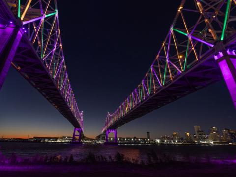 Cresecent City Connection bridge illuminated purple, green, and gold with New Orleans skyline in background.