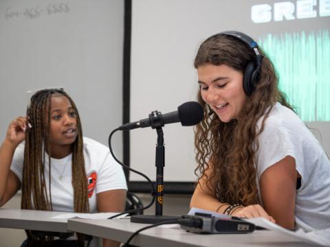 Two pre-college students recording a podcast, one wearing headphones and speaking into a microphone.