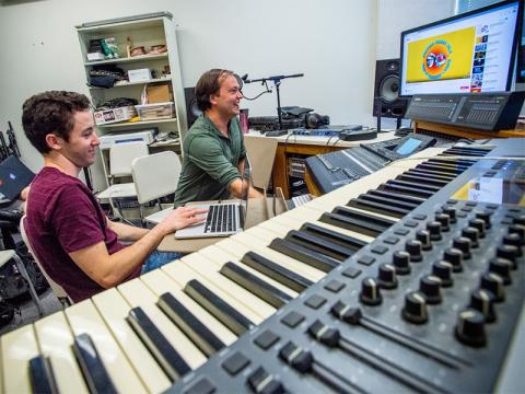 Student and professor smiling while working in a recording studio with a large keyboard.
