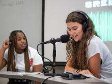Two pre-college students recording a podcast, one wearing headphones and speaking into a microphone.